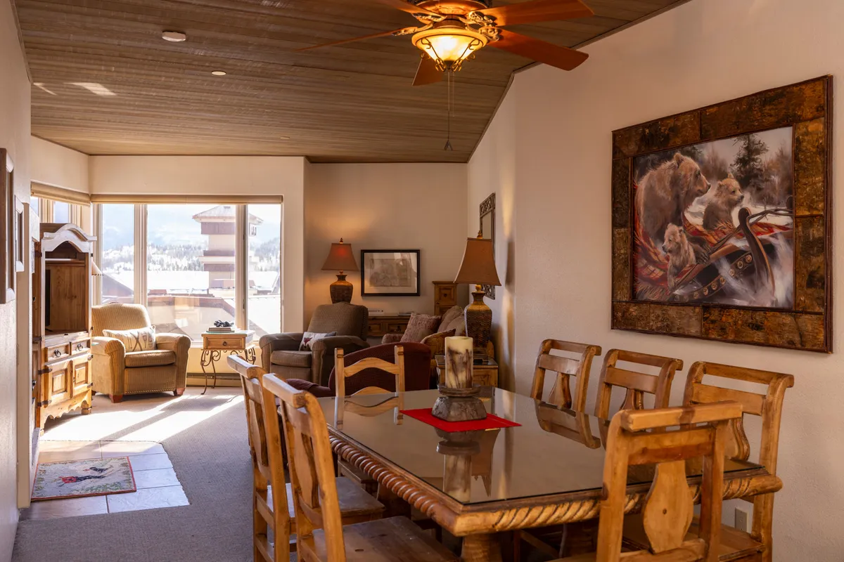 A spacious dining area with a wooden table, surrounded by ample seating. Large windows reveal stunning mountain views, and a bear painting adds a rustic touch. The space feels perfect for shared meals, game nights, and enjoying the changing light over the peaks.