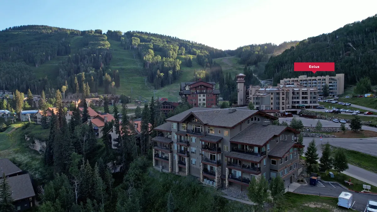 A sweeping exterior shot that brings together the majestic mountain range, golf course, resort buildings, trees, and an expansive parking area in the foreground. The parking area hints at the convenience of reserved, covered parking close to the action, while the stunning landscape underscores year-round recreation.
