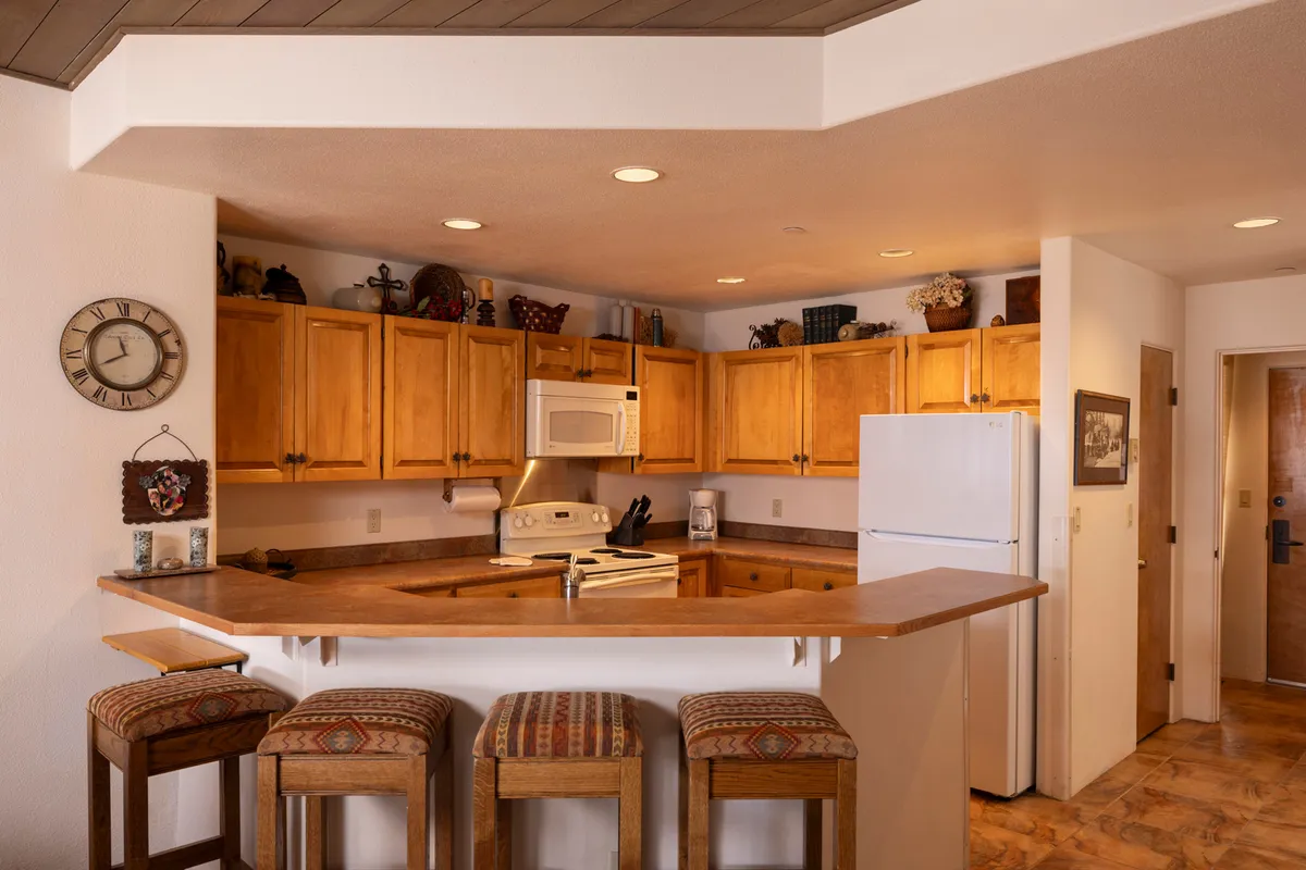 A modern, welcoming kitchen featuring a central island with bar stools, wooden cabinetry, and a classic wall clock. The design is open and oriented for socializing, inviting guests to cook, chat, or enjoy snacks together.