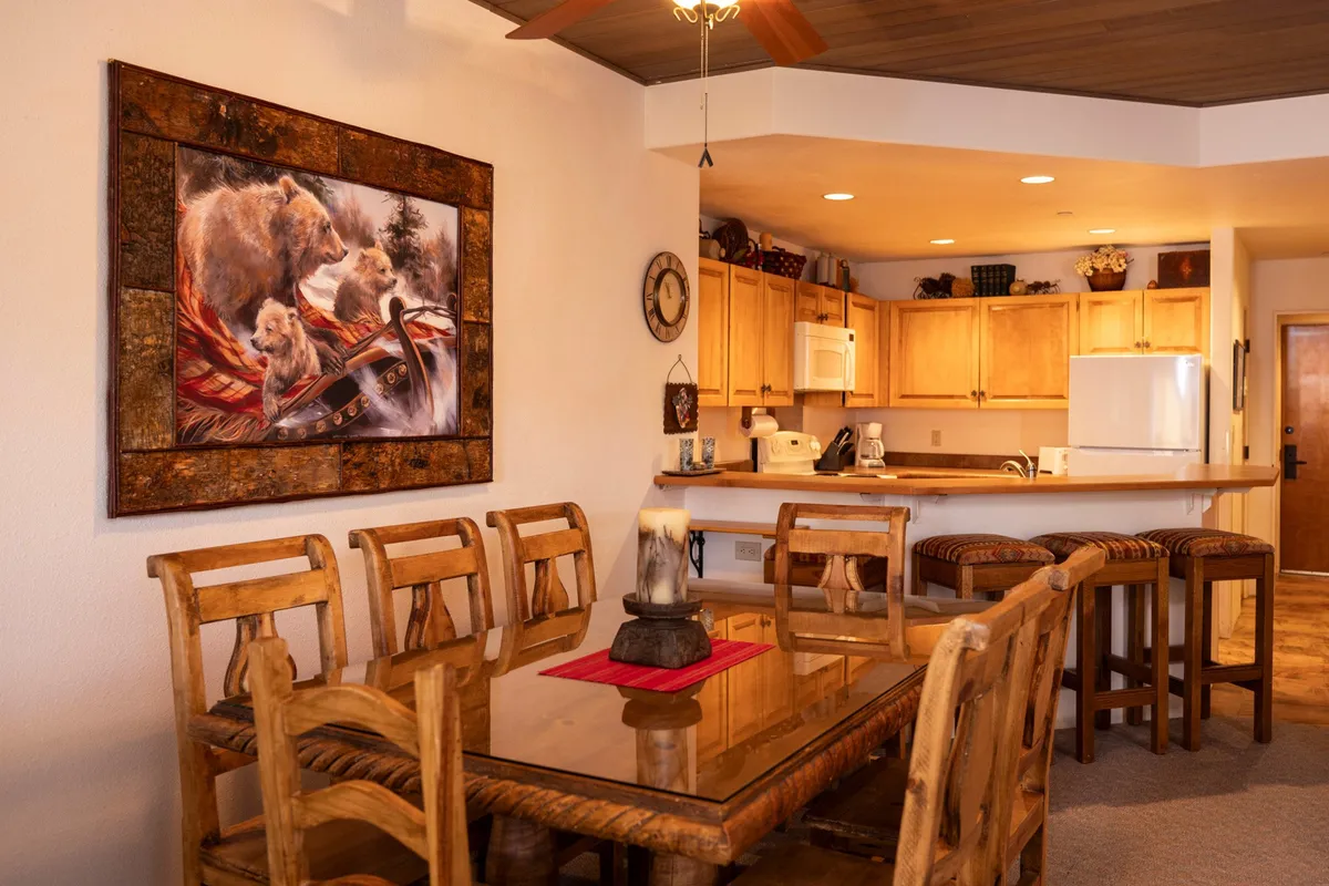 A warm, rustic dining and kitchen area featuring a large wooden dining table, bear-themed artwork, and an open kitchen in the background. The setting hints at shared meals and intimate gatherings after outdoor adventures.