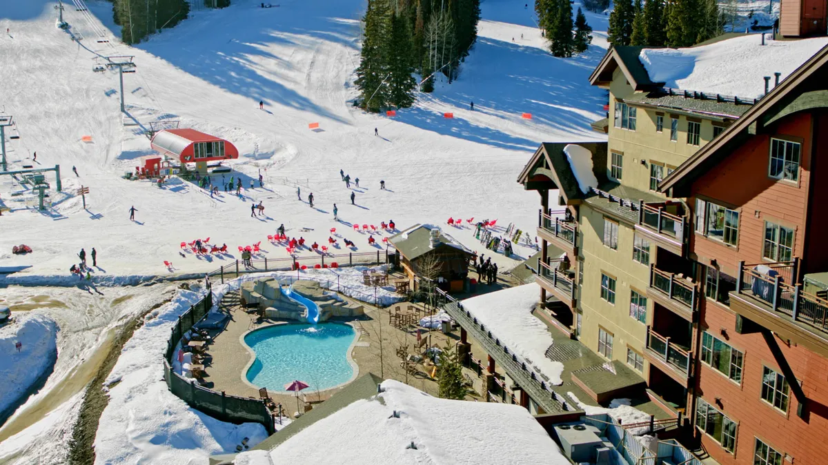 A vibrant exterior view of the property, showcasing the ski slopes, an inviting outdoor pool, the ski lift in close proximity, and the distinctive lodge architecture all set against a snowy landscape. The scene captures people enjoying various activities—skiing, snowboarding, and relaxing by the pool—highlighting the property's direct connection to the mountain environment and recreation.