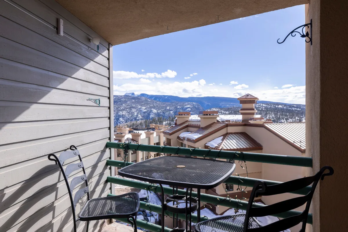 A tranquil private deck with a small bistro table, perfectly positioned to face an expansive mountain view. The balcony railing frames the landscape of peaks and sky, creating an inviting spot for coffee or evening drinks.