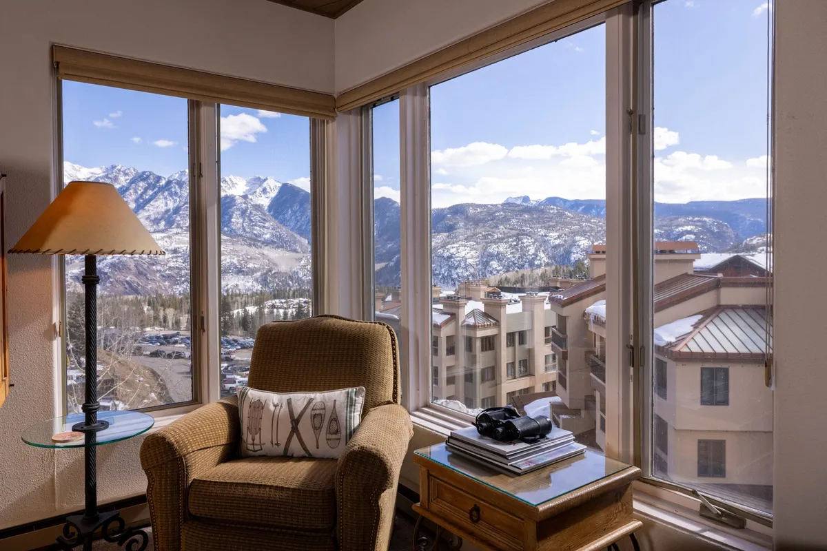 A cozy armchair is positioned beside a table holding books and a camera, all set before expansive floor-to-ceiling windows that frame breathtaking mountain views. Sunlight pours into the inviting living space, offering a tranquil spot for guests to read, relax, or capture the stunning Needles Mountains.