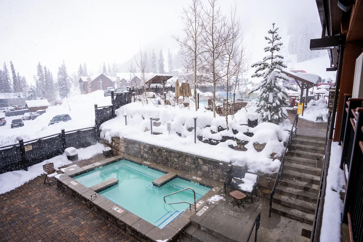This image showcases an outdoor hot tub nestled in a snowy landscape, surrounded by trees and wooden structures. The inviting steam from the hot tub contrasts with the crisp winter scenery, and the overall ambiance is one of relaxation and luxury.