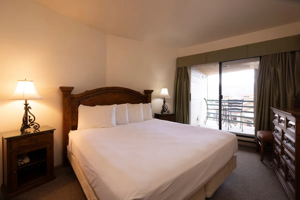 A comfortable bedroom featuring a queen-sized bed with a wooden headboard, bedside lamp, and a door leading to a balcony. The room exudes warmth and tranquility, inviting restful sleep and peaceful mornings.