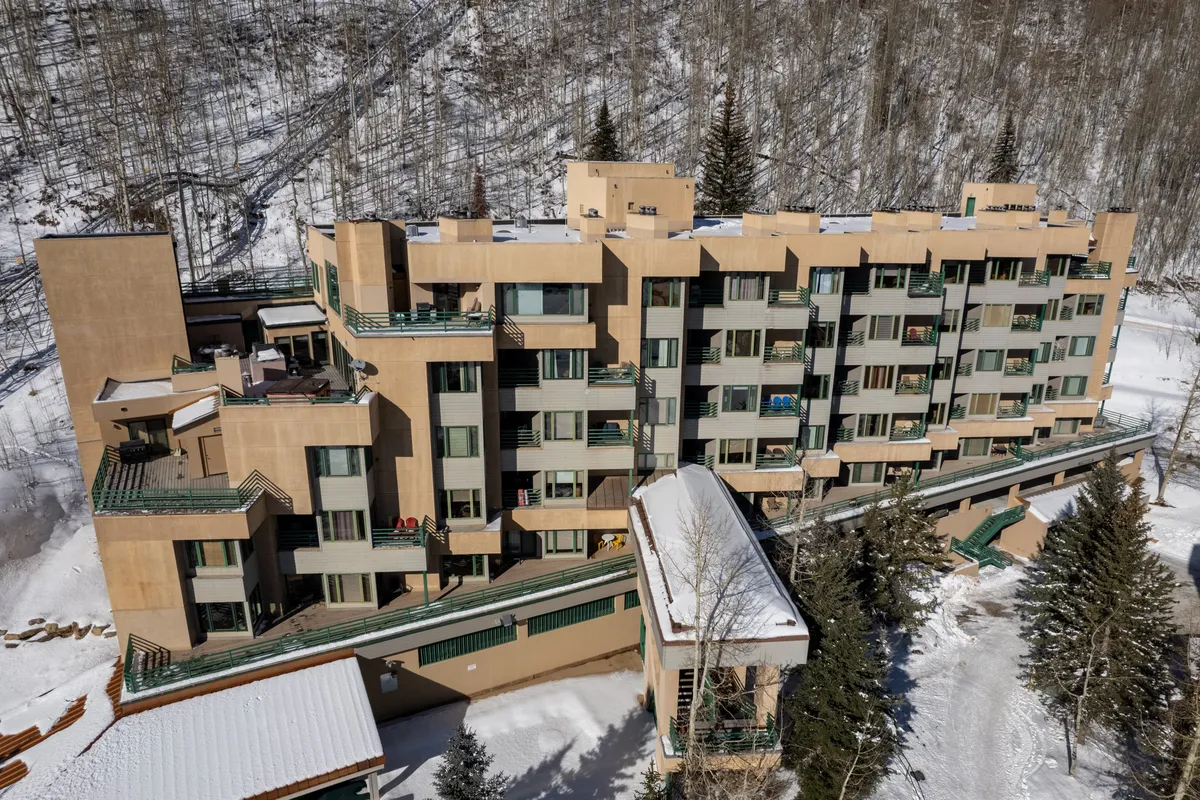 A multi-story building set against a snow-covered landscape, dotted with green balconies and surrounded by trees. The wintery setting showcases both the proximity to the slopes and the inviting nature of the property. The building’s proximity to the snow creates an immediate sense of ski-in/ski-out access, ideal for mountain adventures.