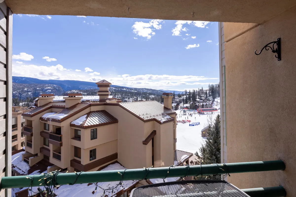 A scenic balcony vantage point overlooking snow-covered ski slopes and distant mountains. The balcony railing frames the view, giving a sense of how close the property is to the action, and inviting guests to imagine stepping out to enjoy the mountain air and watch skiers in action.